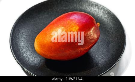 Fresco mango rosso-arancio succoso su un piatto nero isolato su uno sfondo bianco, primo piano Foto Stock