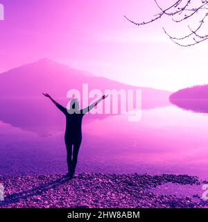 Figura femminile con bracci aperti larghi. Fantastiche vette naturali di montagna riflesse nelle acque del lago. La luce del sole. Foto Stock