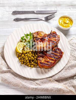 bistecche di carne di maiale alla griglia con piselli verdi bolliti e limone su un piatto bianco su un tavolo di legno, vista verticale dall'alto Foto Stock