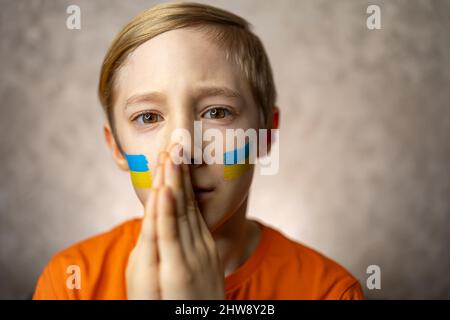 Un bambino contro la guerra, un ragazzo con una bandiera dipinta dell'Ucraina sulle guance ha piegato le mani in un appello per la pace nel mondo Foto Stock