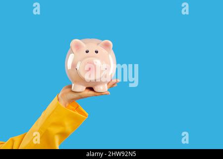 Scatto corto della mano della donna che tiene la banca rosa del piggy sullo sfondo blu dello spazio della copia Foto Stock