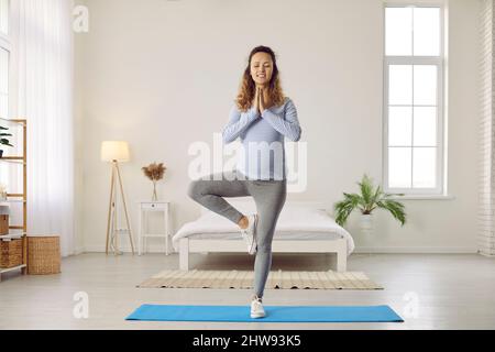 Donna incinta sana felice che fa posa dell'albero mentre pratica gli esercizi di yoga a casa Foto Stock