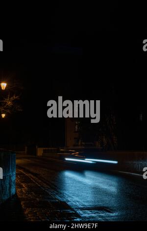 Una foto a lunga esposizione di un'auto in movimento lungo la strada principale di Prestbury, giocando con il contrasto del blu freddo dell'auto e la luce streetlamp. Foto Stock