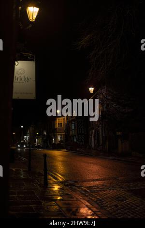 Prestbury High Street in una serata invernale buia e bagnata, con il caldo bagliore delle luci di strada che suonano sui ciottoli. Foto Stock