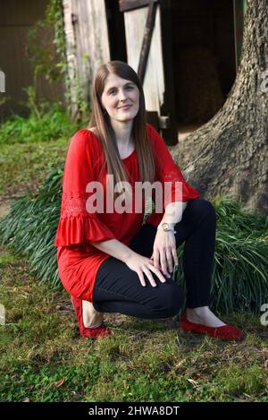 Giovane donna si inginocchia all'esterno di fronte ad un albero e fienile rustico in legno. È smilante e indossa blusa rossa e scarpe rosse. Foto Stock