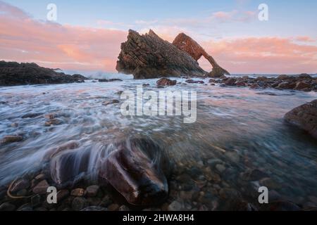 Paesaggio di arco marino naturale e pietre in primo piano coperto di acqua. Famosa formazione rocciosa sulla costa di Moray, Highlands scozzesi, Scozia. Bow Fiddle Rock al tramonto, lunga esposizione. Foto Stock