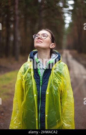 La giovane donna respira aria fresca nella foresta dopo la pioggia, riposa, rilassa, in piedi in un impermeabile giallo, respira ad occhi chiusi Foto Stock