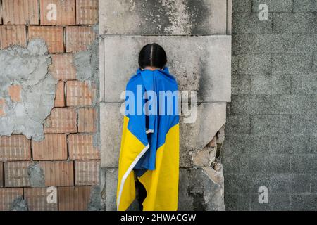 Bambina con bandiera Ucraina davanti ad un muro distrutto dalle bombe. La bambina ondeggia la bandiera nazionale mentre pregava per la pace Foto Stock