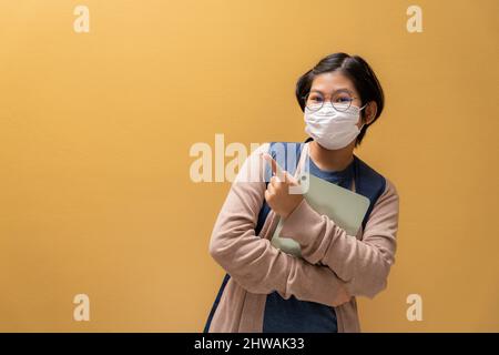 Asian giovane ragazza indossare una maschera protettiva che tiene tablet e libri puntando le mani dito fino a copiare spazio lei indossare giacca maglione e portare zaino shi Foto Stock