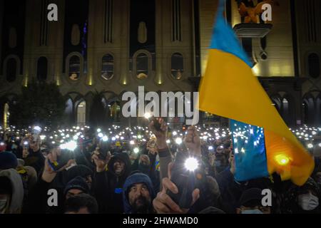 Tbilisi, Georgia. 04th Mar 2022. Una folla di persone tiene in mano le luci del telefono cellulare e una bandiera Ucraina viene sventolata mentre la gente attende il discorso dal vivo del Presidente Volodymyr Zelenskyy. Trentamila persone si sono riunite di fronte all'edificio del parlamento di Tbilisi per ascoltare il discorso dal vivo del presidente ucraino in cui afferma che "l'Ucraina non darà mai il suo territorio alla Russia e l'Ucraina combatterà contro i russi per vincere questa guerra. Voi siete tutti ucraini oggi». Credit: SOPA Images Limited/Alamy Live News Foto Stock