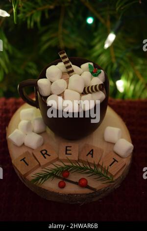 Tazza di cioccolato piena di marshmallows bianchi e bastoni di caramelle su legno rotondo con la parola 'trattare' con albero in background - vacanza still life. Foto Stock