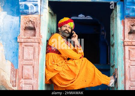 Felice uomo maturo indiano del nord indossando abiti colorati e turbante parlare su smartphone, sorridente tradizionale maschio chiamata su android smart mobile pho Foto Stock