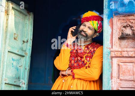 Felice uomo maturo indiano del nord indossando abiti colorati e turbante parlare su smartphone, sorridente tradizionale maschio chiamata su android smart mobile pho Foto Stock