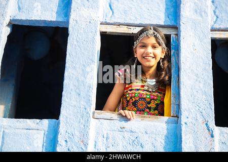 Bambino ragazza indiana piccola felice che indossa tradizionale rajasthani colorato vestito e gioielleria che guarda fuori dalla finestra. Foto Stock