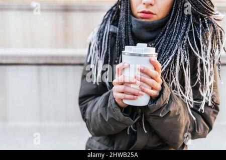 Taglia la donna irriconoscibile in un outerwear caldo con dreadlock che tiene thermos bianchi con caffè caldo nelle mani mentre si alza in strada Foto Stock