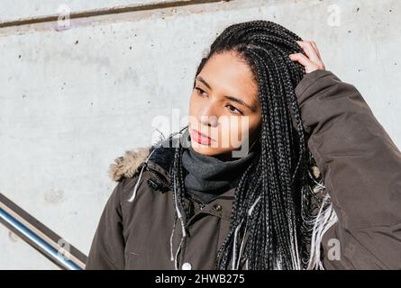 Hipster femminile freddo con trecce lunghe che regolano i capelli mentre si trova in piedi in abiti caldi in strada nelle giornate di sole e guardando lontano Foto Stock