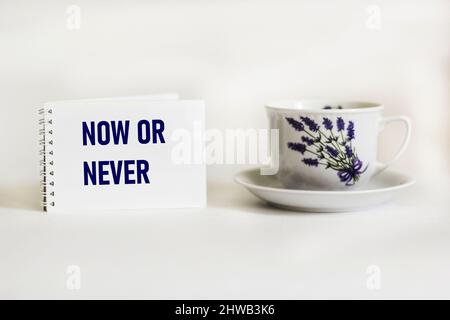Citazione di ispirazione ora o mai su un blocco note con una tazza d'epoca bella e sfondo bianco .tipografia motivazionale. Foto Stock