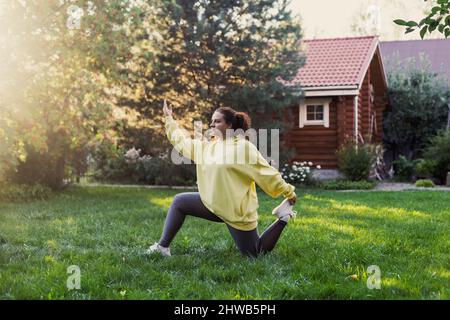 Giovane donna che indossa abiti sportivi facendo esercizi di yoga sul cortile sul prato verde con casa di campagna in legno e alti alberi sullo sfondo. Corpo Foto Stock