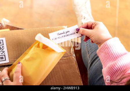 Alto angolo di raccolto anonimo mittente femmina imballaggio ordine in busta e mettere nota grazie per l'acquisto prima di spedizione in scatola Foto Stock