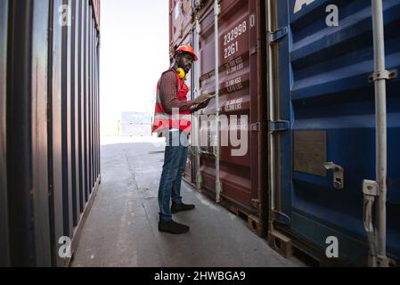 Lavoratore maschio afroamericano che tiene tavoletta e controlla il carico di contenitori di merci da carico Cargo nave per l'esportazione di importazione. Foto Stock