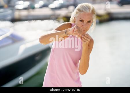 Donna matura che fa boxe ombra all'aperto. Donna anziana che fa sport in un porto costiero Foto Stock