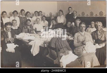 Riunione di cucitura del Monte Gould, Plymouth, 28 agosto 1917: Gruppo di (soprattutto) donne impegnate nella cucitura a mano e nella cucitura a macchina. Macchine da cucire e pile di tessuto sono su un tavolo da lavoro con donne sedute intorno ad esso. Tre uomini (uno un clergyman) e sette donne sono in piedi sullo sfondo Foto Stock