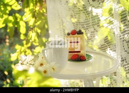 Bella tazza di caffè in porcellana e fetta di torta su un tavolo di vetro decorato con fiori bianchi nel giardino estivo alla luce del sole. Spazio di copia. Concetto di bevande estive. Foto Stock