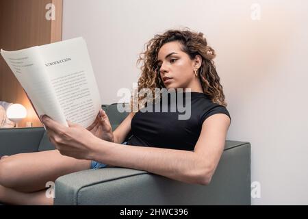 ritratto di giovane donna caucasica in abiti casual seduta sul divano a casa girando la pagina del libro che sta leggendo, stile di vita e rea Foto Stock