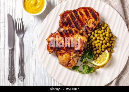 bistecche di carne di maiale alla griglia con piselli verdi bolliti e limone su un piatto bianco su un tavolo di legno con mostarda e posate, piatto Foto Stock