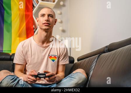 Calvo queer attivista caucasico usando gamepad e cercando di vincere una partita. Bandiera arcobaleno sullo sfondo. Foto di alta qualità Foto Stock