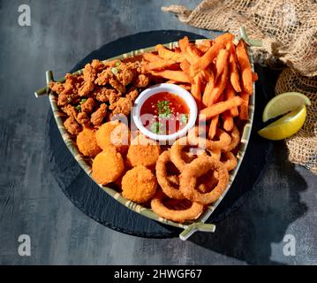 Piatto Snacker con anello di cipolla, patatine fritte, capesante dorate, salsa al peperoncino in un piatto vista dall'alto su sfondo scuro Foto Stock