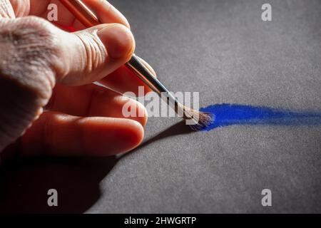 Arte e tema artigianale che mostra una mano di pittura con pennello e vernice blu su terreno grigio Foto Stock