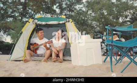 Giovane coppia amorevole grazioso mentre accampamento suona la chitarra mentre si siede alla tenda di campeggio sulla spiaggia in vacanza tempo al tramonto, giovane gruppo asiatico Foto Stock