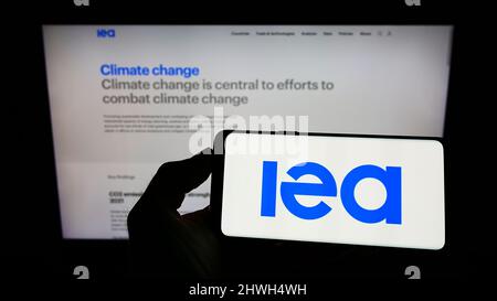 Persona che tiene il cellulare con il logo dell'organizzazione Agenzia Internazionale dell'energia (IEA) sullo schermo di fronte alla pagina web. Mettere a fuoco sul display del telefono. Foto Stock