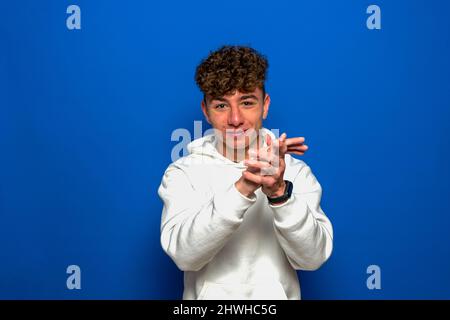 Giovane bruna bella uomo con capelli ricci indossando una felpa bianca che stringe le mani su sfondo blu e che stringe felice e gioioso, sorridente Foto Stock