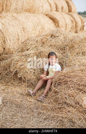 Bella ragazza giovane seduta in mucchio di fieno sparso sorridente con occhi di pensiero sly guardando via e tenendo fieno in mano indossando la Sundress. Divertitevi Foto Stock