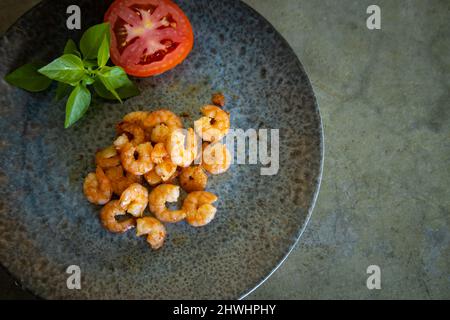 Deliziosi gamberi appena cotti su un piatto grigio accanto a un pomodoro e basilico. Spazio libero per scrivere. Sfondo grigio neutro. Foto Stock