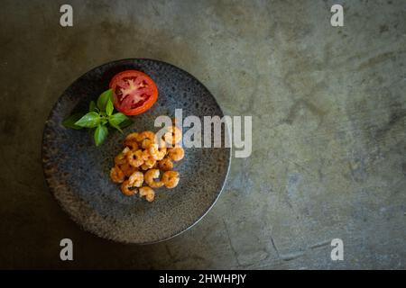 Deliziosi gamberi appena cotti su un piatto grigio accanto a un pomodoro e basilico. Spazio libero per scrivere. Sfondo grigio neutro. Foto Stock