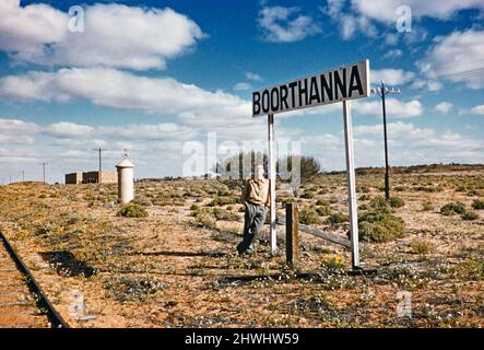 Giovane uomo appoggiato contro il cartello della stazione ferroviaria, Boorthanna, South Australia, Australia 1956 un lato sulla vecchia linea ferroviaria di Ghan ufficialmente la sezione di Port Augusta-Oodnadatta della ferrovia dell'Australia Centrale che ha chiuso il 1960 Foto Stock