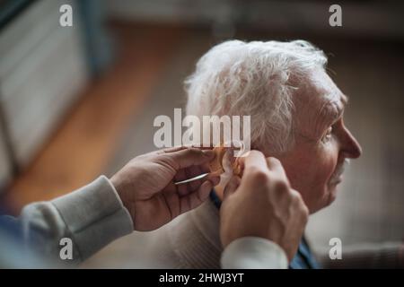 Primo piano della mano dell'operatore sanitario inserendo l'apparecchio acustico nell'orecchio dell'uomo anziano. Foto Stock