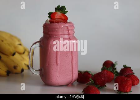 Frullato a base di fragole fresche, banana e latte di mandorle. Servito in vasetto di mason. Girato su sfondo bianco con banane e fragole intorno. Foto Stock