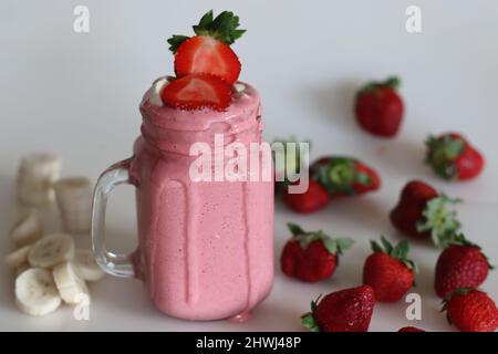 Frullato a base di fragole fresche, banana e latte di mandorle. Servito in vasetto di mason. Girato su sfondo bianco con banane e fragole intorno. Foto Stock