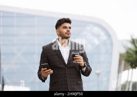 Bell'uomo d'affari indiano in abbigliamento formale, tenendo una tazza di caffè guardando via. Il giovane uomo arabo pensieroso si erge fuori dall'edificio moderno e beve Foto Stock