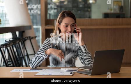 Immagine dell'affascinante donna asiatica che parla con lo smartphone e lavora su un computer portatile, mentre si siede al tavolo al coperto. Foto Stock