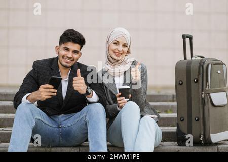 Due uomini d'affari multirazziali seduti alle scale con borsa da viaggio e guardando la macchina fotografica che mostra i pollici in su. Diversi colleghi pronti per il viaggio, seduti con i telefoni e il caffè in strair all'aperto Foto Stock