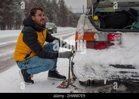 Il giovane uomo sta installando catene da neve su un'auto d'epoca su strade innevate e ghiacciate. Uomo che lotta per ottenere le catene da neve auto su pneumatici invernali. Foto Stock