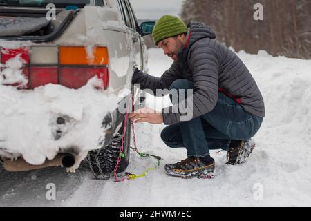 Il giovane uomo sta installando catene da neve su un'auto d'epoca su strade innevate e ghiacciate. Uomo che lotta per ottenere le catene da neve auto su pneumatici invernali. Foto Stock