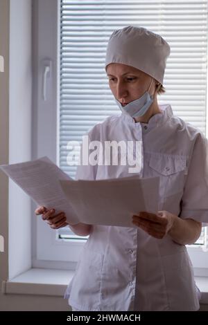 Una dottoressa con una maschera sul viso si trova vicino alla finestra e guarda attraverso i documenti. Foto Stock