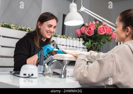 Giovane manicurista indossare guanti con gioia guardando un cliente mentre si rimuove un vecchio manicure da una giovane donna con una lima unghie in un salone di bellezza. Foto Stock