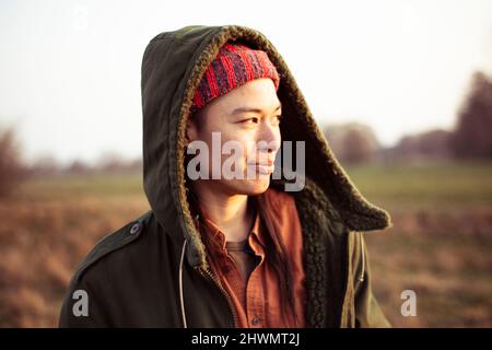 Persona non binaria sorride al sole del pomeriggio all'aperto in campo con il cappuccio Foto Stock